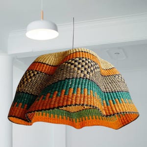 Könnte beinhalten: Eine gewebte Pendelleuchte mit farbenfrohem Design. Der Schirm hat horizontale Bänder in Orange, Grün, Schwarz und Beige. Im Hintergrund ist eine weiße, runde Deckenleuchte zu sehen.