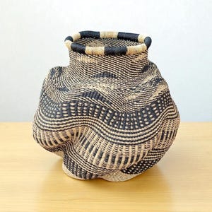 Puede incluir: Una cesta tejida a mano con una forma orgánica única. La cesta presenta una combinación de fibras tejidas beige natural y negras, creando un patrón de rayas. El borde está acabado con un diseño de tablero de ajedrez negro y beige.