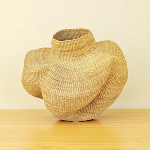 May include: A woven basket with a unique, sculptural form. The basket is made of natural-colored fibers, with a cylindrical neck and a body that curves and twists. The basket is displayed on a wooden surface.