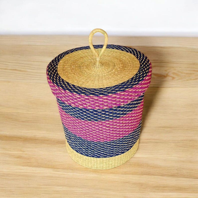 May include: A handwoven storage basket with a lid. The basket has horizontal stripes in pink, navy blue, and natural beige. The lid has a loop handle. The basket is made of natural fibres.