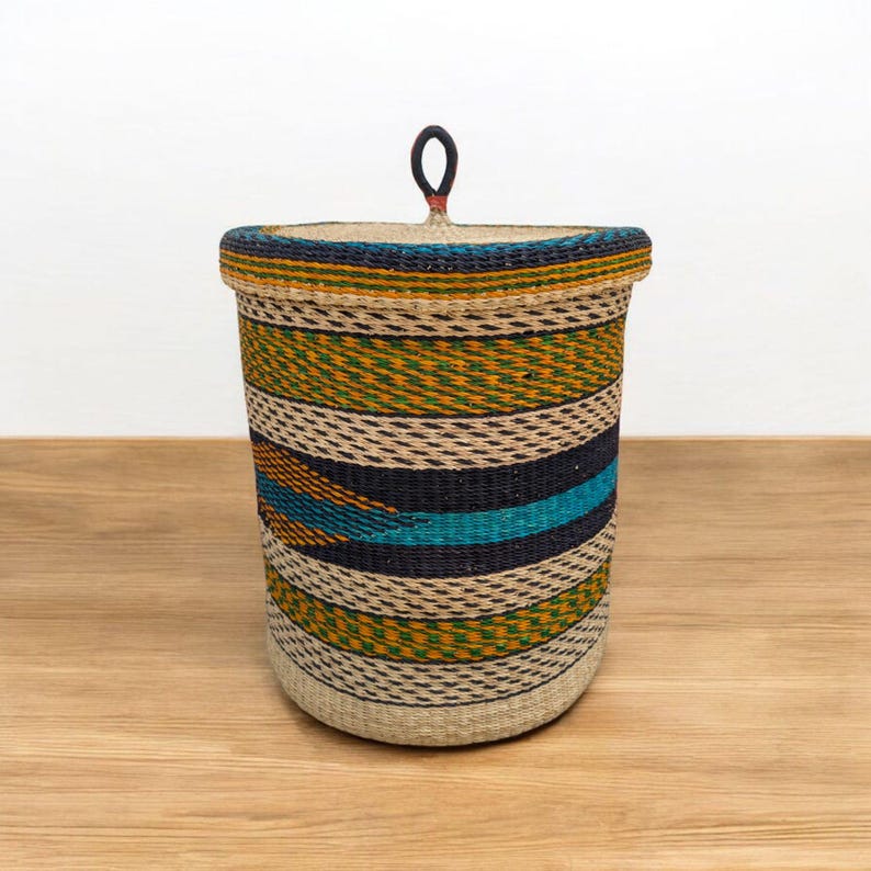 May include: A round woven storage basket with a lid and a handle. The basket has horizontal stripes in beige, black, teal, and yellow-green. The lid has a small, dark red handle. The basket is made of natural fibres.