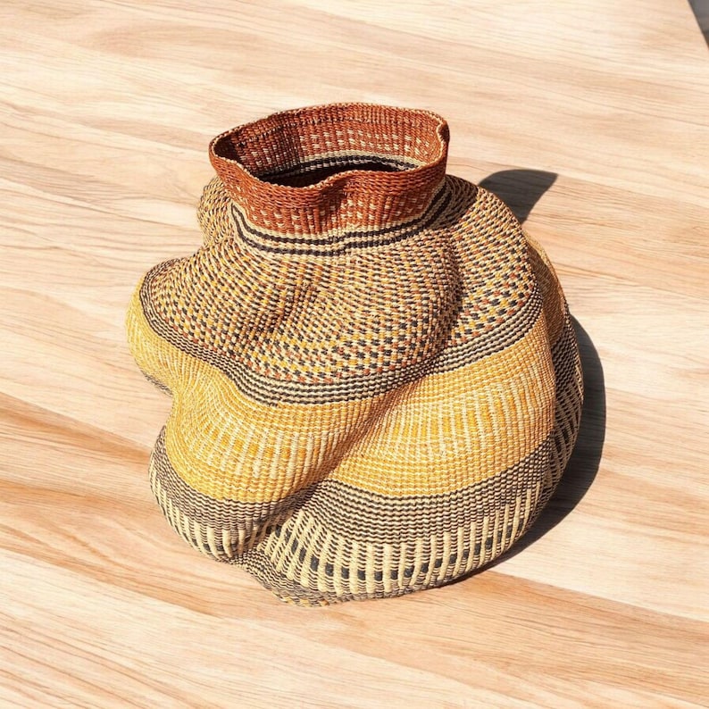 May include: Handwoven basket with a flared rim and a bulbous body. The basket features alternating bands of yellow, black, and natural fiber tones. The rim is a darker brown color, adding contrast to the overall design.