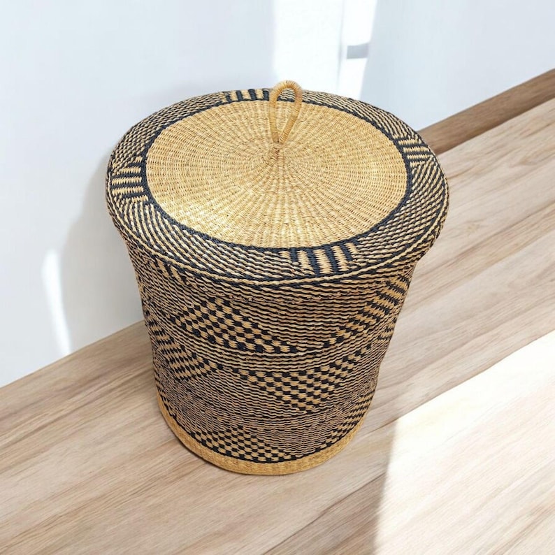 May include: A round, woven storage basket with a lid. The basket is cylindrical with a natural tan colour and a black geometric pattern. The lid has a loop handle. The basket is made of natural fibres.