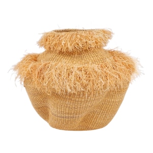 Könnte beinhalten: Ein gewebter Korb in hellem Beige mit einer einzigartigen Form. Er hat einen ausgestellten Boden und eine schmalere Oberseite, mit einem Fransenbesatz aus strohartigem Material um die Mitte und die obere Öffnung. Der Korb ist wahrscheinlich handgefertigt.