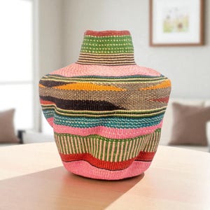 Puede incluir: Un jarrón decorativo tejido con un cuerpo ancho y un cuello estrecho. El jarrón presenta rayas horizontales en varios colores, incluyendo rosa, verde, azul, amarillo y negro. El jarrón está sobre una mesa de madera clara.