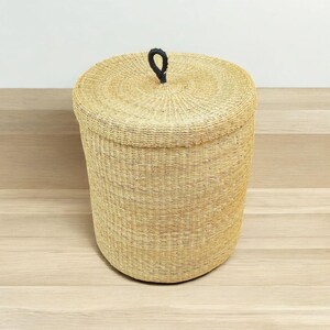 May include: A round, woven basket with a lid. The basket is made of natural-coloured fibres and has a black handle on the lid. The basket is cylindrical in shape and sits on a light-coloured wooden surface. The basket is likely used for storage or laundry.