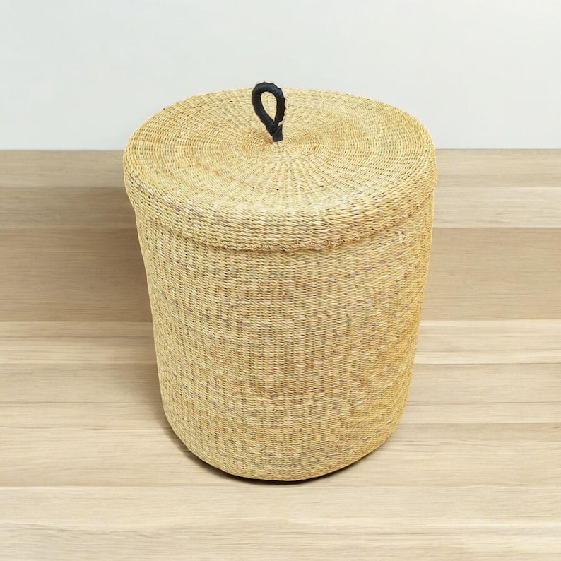 May include: A round, woven basket with a lid. The basket is made of natural-coloured fibres and has a black handle on the lid. The basket is cylindrical in shape and sits on a light-coloured wooden surface. The basket is likely used for storage or laundry.