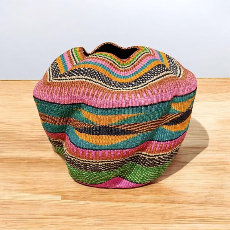 May include: A vibrantly colored woven basket with a unique, wavy shape. The basket features a mix of pink, green, orange, and black stripes. The interior is dark, and the basket sits on a wooden surface.