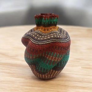 May include: A handwoven basket with a unique, asymmetrical shape. The basket features a vibrant color palette of red, green, brown, and yellow, arranged in horizontal and diagonal stripes. The top of the basket has a ruffled edge.