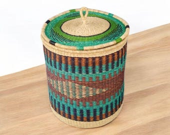 May include: A woven storage basket with a lid. The basket is cylindrical with a natural beige base and lid. The body features a pattern of teal, brown, and navy blue horizontal stripes. The lid has a green and beige circular design.