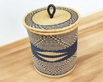 May include: A cylindrical woven basket with a lid. The basket is made of natural and navy blue fibers, with a repeating geometric pattern. The lid has a black loop handle.