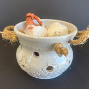 May include: A white ceramic garlic keeper with a rope handle. The keeper is filled with garlic and an onion. The keeper has decorative cutouts and is on a dark surface.