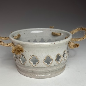May include: A speckled, off-white ceramic bowl with a decorative quatrefoil pattern and rope handles. The bowl has a wide, shallow shape and a slightly textured surface. The rope handles are attached to the sides of the bowl.