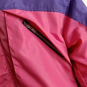 May include: Close-up of a pink and purple jacket. The jacket features a pink body with a purple upper section. A black zipper pocket is visible on the front. The jacket appears to be made of a smooth, possibly nylon, material.