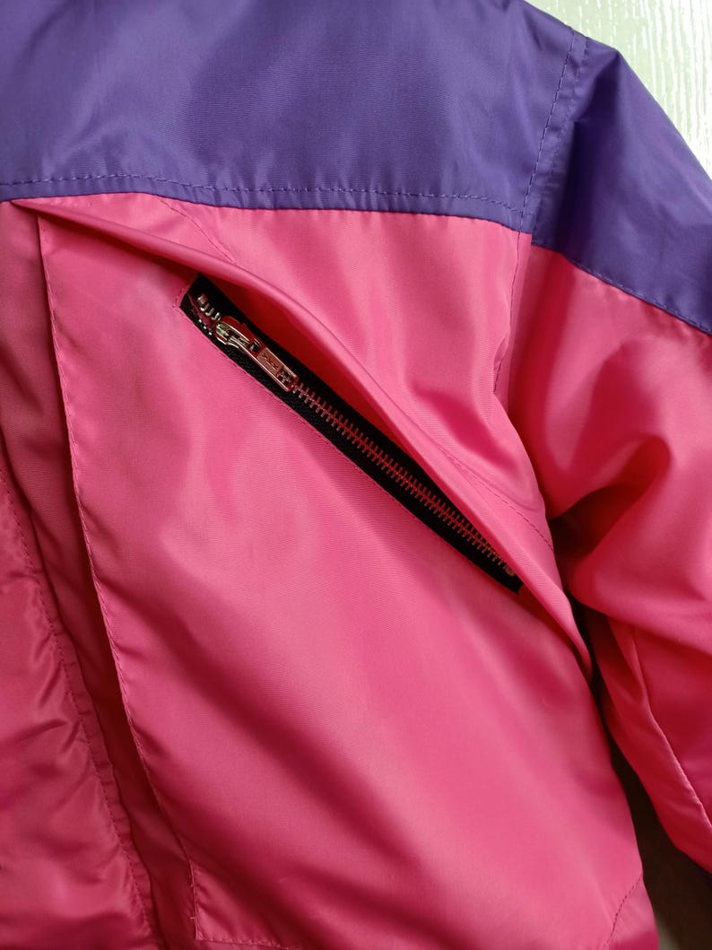 May include: Close-up of a pink and purple jacket. The jacket features a pink body with a purple upper section. A black zipper pocket is visible on the front. The jacket appears to be made of a smooth, possibly nylon, material.