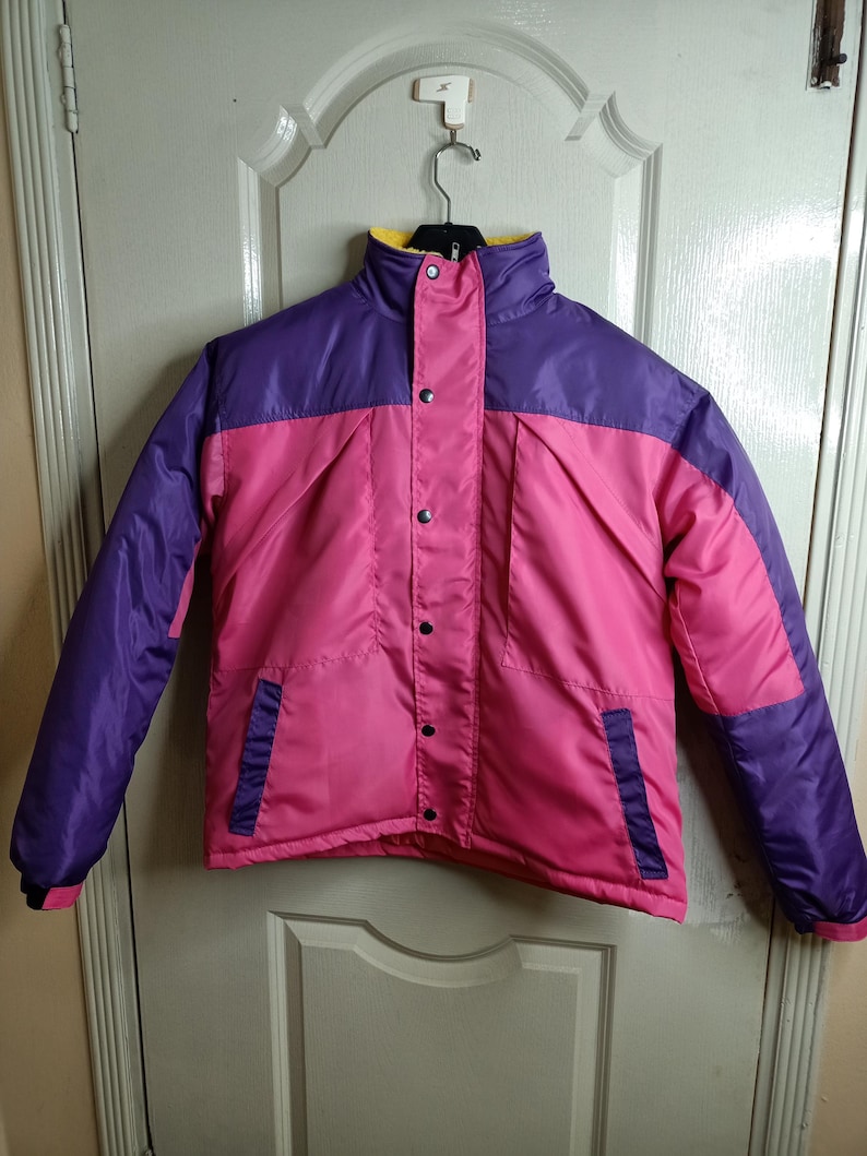 May include: A color-blocked jacket in pink, purple, and yellow. The jacket has a high collar, snap closures, and two front pockets. The sleeves and upper chest are purple, while the body is pink.