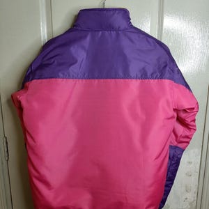 May include: A vibrant, color-blocked jacket in shades of purple and pink. The jacket features a high collar and a smooth, shiny fabric. The back view showcases the color-blocking design, with a horizontal division between the purple upper section and the pink lower section.