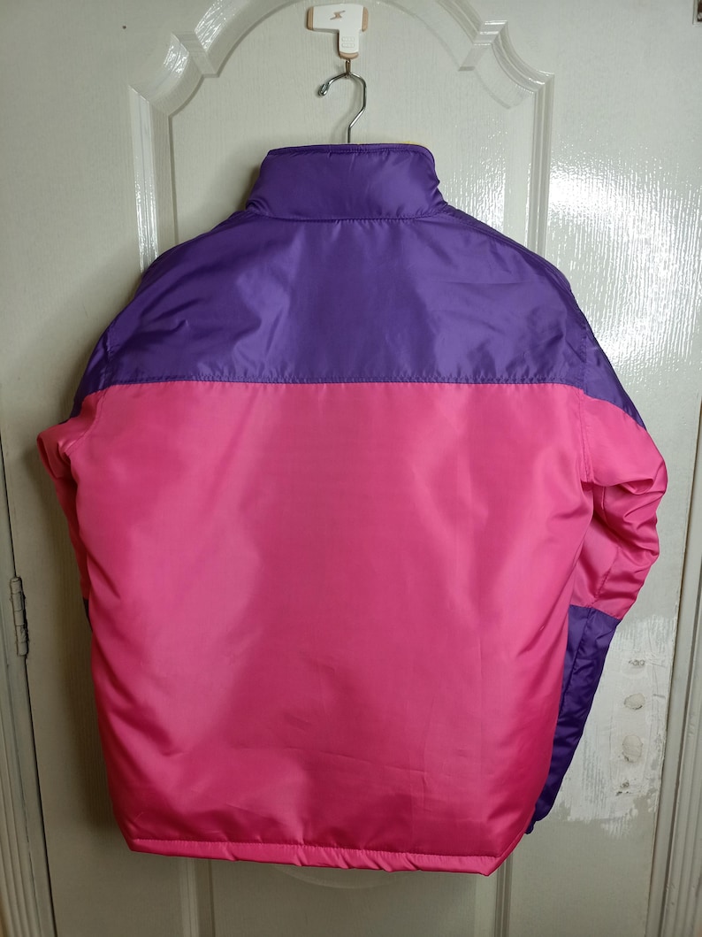 May include: A vibrant, color-blocked jacket in shades of purple and pink. The jacket features a high collar and a smooth, shiny fabric. The back view showcases the color-blocking design, with a horizontal division between the purple upper section and the pink lower section.