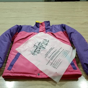 May include: A pink, purple, and yellow jacket with a high collar and snap closures. The jacket has purple sleeves and a pink body, with a yellow collar. A clear plastic bag with text is on top of the jacket.