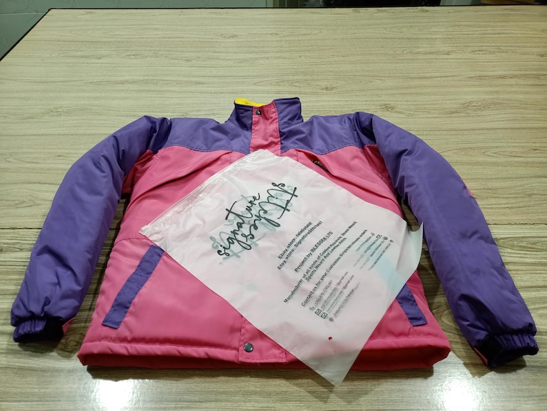 May include: A pink, purple, and yellow jacket with a high collar and snap closures. The jacket has purple sleeves and a pink body, with a yellow collar. A clear plastic bag with text is on top of the jacket.
