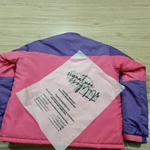 May include: A pink and purple jacket with a matching pink plastic bag on top. The jacket has a pink body with purple shoulders and sleeves. The bag has the words "signature stitches" printed on it.