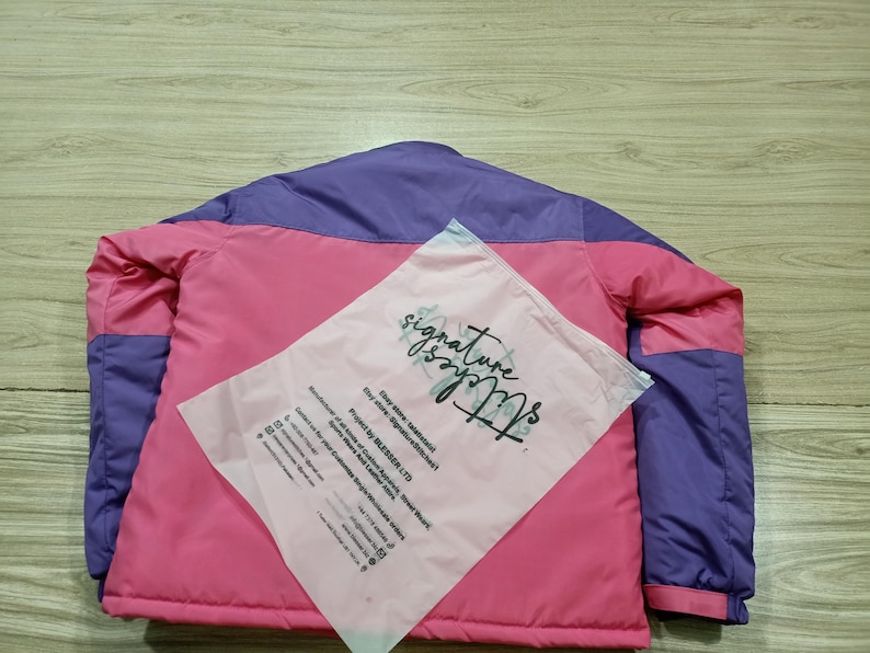 May include: A pink and purple jacket with a matching pink plastic bag on top. The jacket has a pink body with purple shoulders and sleeves. The bag has the words "signature stitches" printed on it.