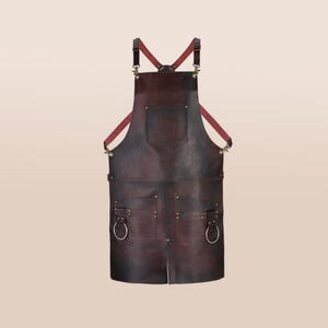 Könnte beinhalten: Eine dunkelbraune Lederschürze mit Kreuzrücken-Design. Die Schürze hat zwei große Taschen, eine Brusttasche und Metallring-Akzente. Die Träger sind bordeauxfarben und die Schürze ist für die Küche oder Werkstatt konzipiert.