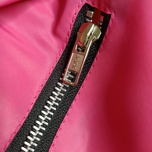 May include: Close-up of a bright pink fabric with a black zipper. The zipper is silver and has the YKK brand name on the pull tab. The fabric is smooth and appears to be a jacket or coat.