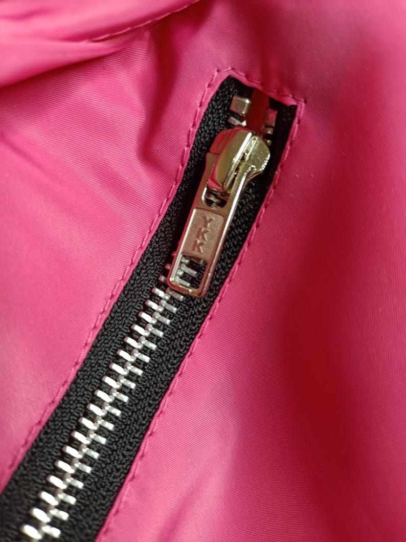 May include: Close-up of a bright pink fabric with a black zipper. The zipper is silver and has the YKK brand name on the pull tab. The fabric is smooth and appears to be a jacket or coat.