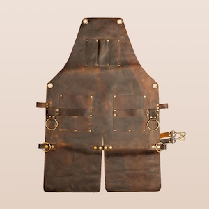 Könnte beinhalten: Eine braune Lederschürze mit mehreren Taschen und Messingbeschlägen. Die Schürze hat zwei große Taschen, zwei kleinere Taschen und Messingringe zum Befestigen von Werkzeugen. Die Schürze hat ein geteiltes Design am unteren Ende und wird mit Riemen und Schnallen befestigt.