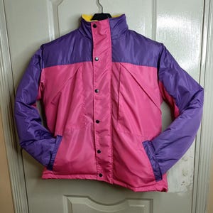 May include: A colorful, vintage jacket in pink, purple, and yellow. The jacket has a high collar, snap closures, and two front pockets. The sleeves and upper yoke are purple, while the body is pink. The collar lining is yellow.