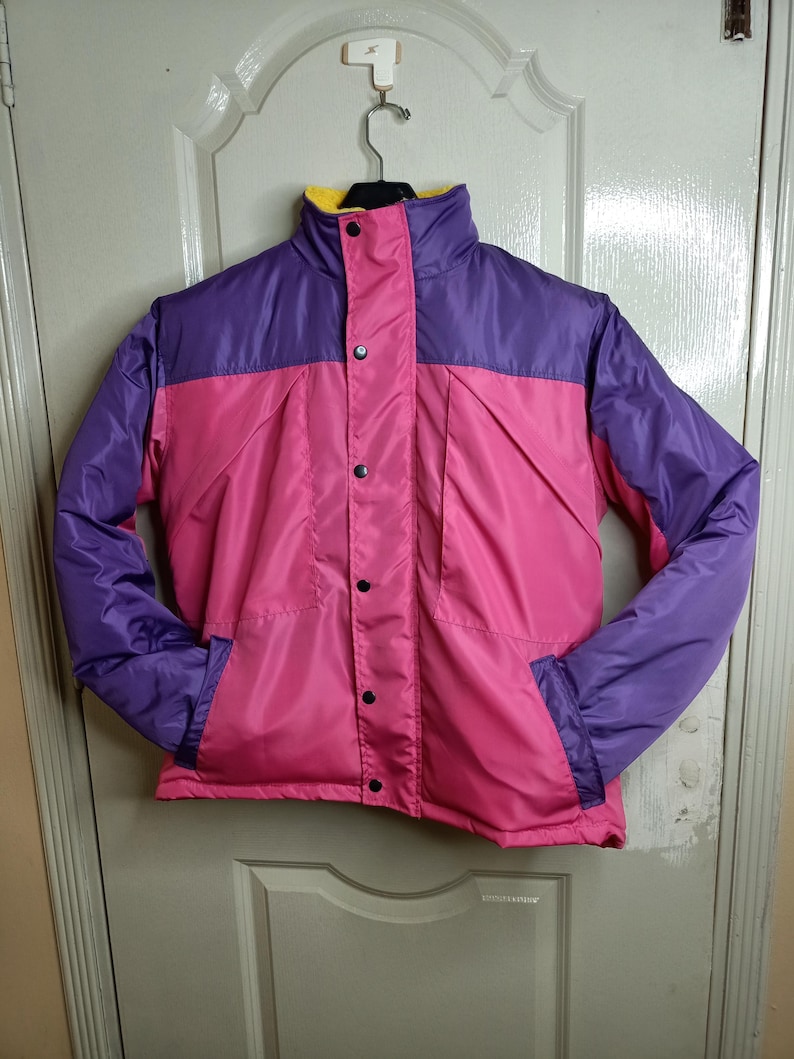 May include: A colorful, vintage jacket in pink, purple, and yellow. The jacket has a high collar, snap closures, and two front pockets. The sleeves and upper yoke are purple, while the body is pink. The collar lining is yellow.