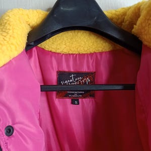 May include: A small, size S, jacket with a bright pink interior and a yellow, fuzzy collar. The jacket is hanging on a black hanger. The label reads "signature" and "MANUFACTURED BY BLESSER LTD."