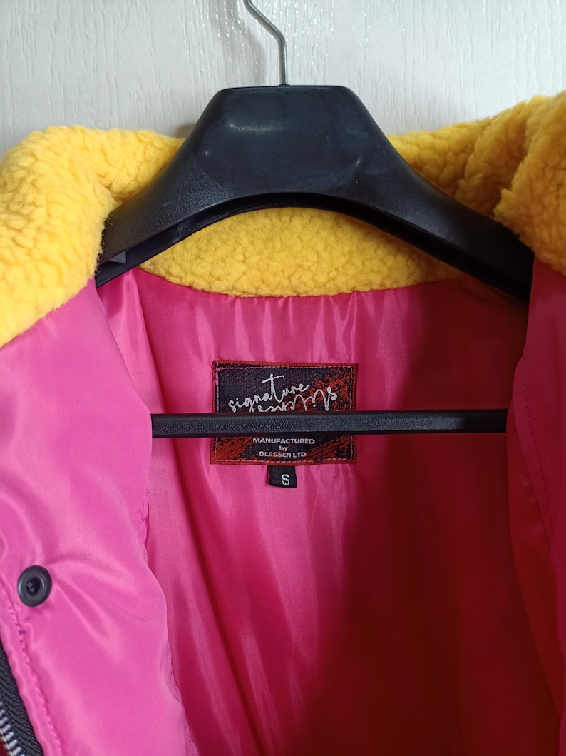 May include: A small, size S, jacket with a bright pink interior and a yellow, fuzzy collar. The jacket is hanging on a black hanger. The label reads "signature" and "MANUFACTURED BY BLESSER LTD."