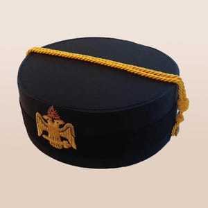 Puede incluir: Sombrero negro con un emblema de águila bordado en oro y un detalle de cuerda amarilla. El sombrero tiene forma cilíndrica y una parte superior plana. El emblema está centrado en la parte delantera.
