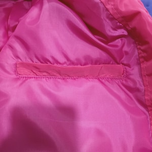 May include: Close-up of the inside of a bright pink jacket. The lining is a smooth, shiny pink fabric with a rectangular pocket. The jacket's exterior is a darker pink and purple.