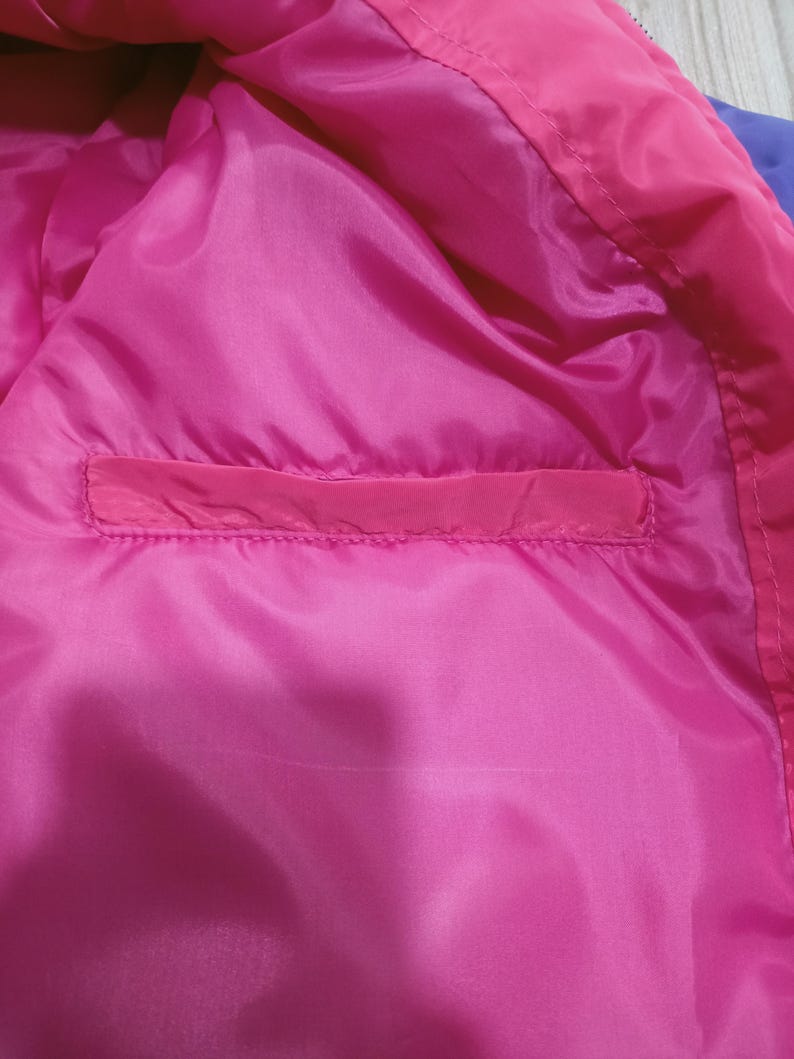 May include: Close-up of the inside of a bright pink jacket. The lining is a smooth, shiny pink fabric with a rectangular pocket. The jacket's exterior is a darker pink and purple.