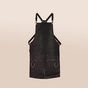 May include: Black leather apron with adjustable straps and multiple pockets. Features brass-colored rivets and metal rings. The apron has a cross-back design and is suitable for various activities.
