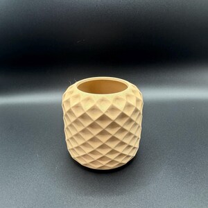 May include: A cylindrical, light brown vase with a geometric diamond pattern. The vase has a wide opening and is made of a natural-looking material. The design gives the vase a textured appearance. The vase is suitable for holding small plants or decorative items.
