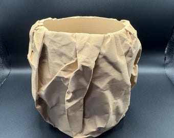 May include: A beige, fabric-covered decorative bowl with a textured, pleated design. The bowl has a wide opening and a rounded base. The fabric appears to be a light, neutral color, and the bowl is set against a dark background.