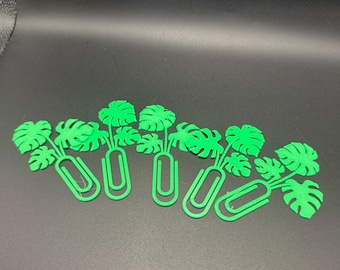 May include: Five green paper clips, each adorned with a stylized monstera leaf design. The clips are arranged on a dark surface, showcasing the bright green color of the decorative elements.