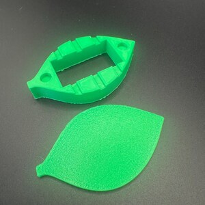 May include: A bright green, leaf-shaped 3D-printed object, with a matching leaf-shaped piece next to it. The top piece has a rectangular opening and two holes. The bottom piece is a solid leaf shape. The objects are on a dark surface.