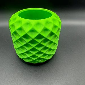 May include: A bright green, 3D-printed vase with a geometric diamond pattern. The vase has a cylindrical shape with a wide opening at the top. The textured surface catches the light, creating shadows and highlights. The vase is suitable for holding flowers or as a decorative item.
