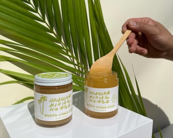 Pode incluir: Dois frascos de vidro de mel com rótulos brancos que dizem "Hawaiian Kiawe Honey" e "North Shore Bees". Uma colher de pau retira mel de um frasco. O fundo apresenta uma grande folha de palmeira verde.