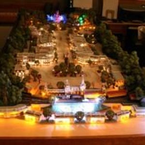 May include: A detailed miniature model of a town square, illuminated with warm yellow and colorful lights. The model features buildings, trees, and a central fountain, creating a vibrant and inviting scene. The model is displayed on a wooden surface.
