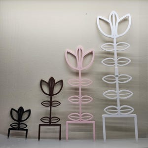May include: Four plant trellises of varying heights arranged in a row. The trellises are black, dark brown, light pink, and white, each featuring a central stem with leaf-shaped cutouts and a pointed top, supported by two prongs at the base.
