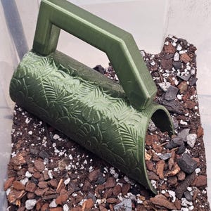 May include: A dark olive green soil scoop featuring a textured surface with an embossed pattern of various tropical leaves. The scoop has a sturdy, angular handle and is filled with a mixture of dark potting soil, wood chips, and perlite.