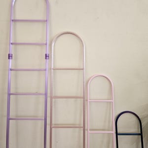 May include: Four arched plant trellises of varying heights arranged in a row. The tallest is purple with a central connector, the second is a textured light pink, the third is a solid light pink, and the shortest is a dark blue gradient.