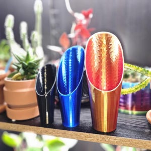 May include: Three cylindrical scoops with angled openings, arranged in a row. The leftmost scoop is black, the middle scoop is metallic blue, and the rightmost scoop features a gradient of red, orange, and yellow with a textured, faceted surface.