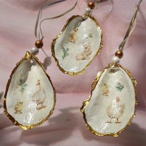 Puede incluir: Tres adornos decorativos de concha de ostra con bordes dorados y cintas para colgar. Cada concha presenta una escena de animales de granja pintada con un pato, un pollito y una gallina. Las conchas están adornadas con cuentas.